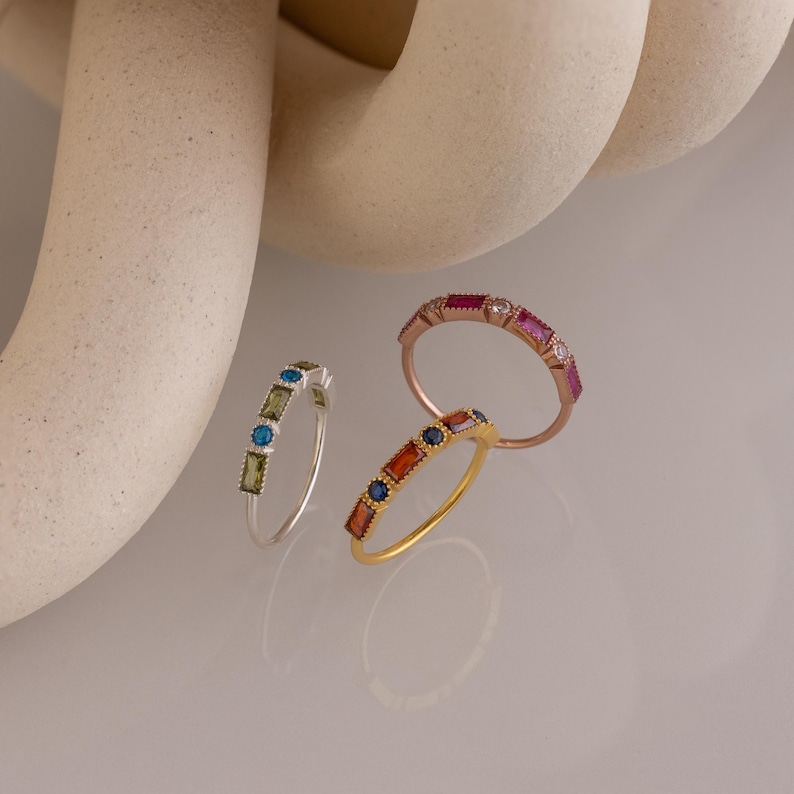 May include: Three gemstone rings in silver, gold, and rose gold. The silver ring has green and blue stones. The gold ring has orange and blue stones. The rose gold ring has pink and white stones. The rings are displayed on a reflective surface.