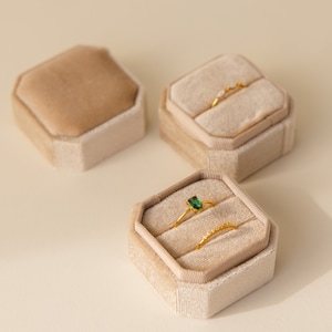 May include: Three velvet ring boxes in beige, with two rings inside one of the boxes. One ring has a green gemstone, and the other ring has small white stones.