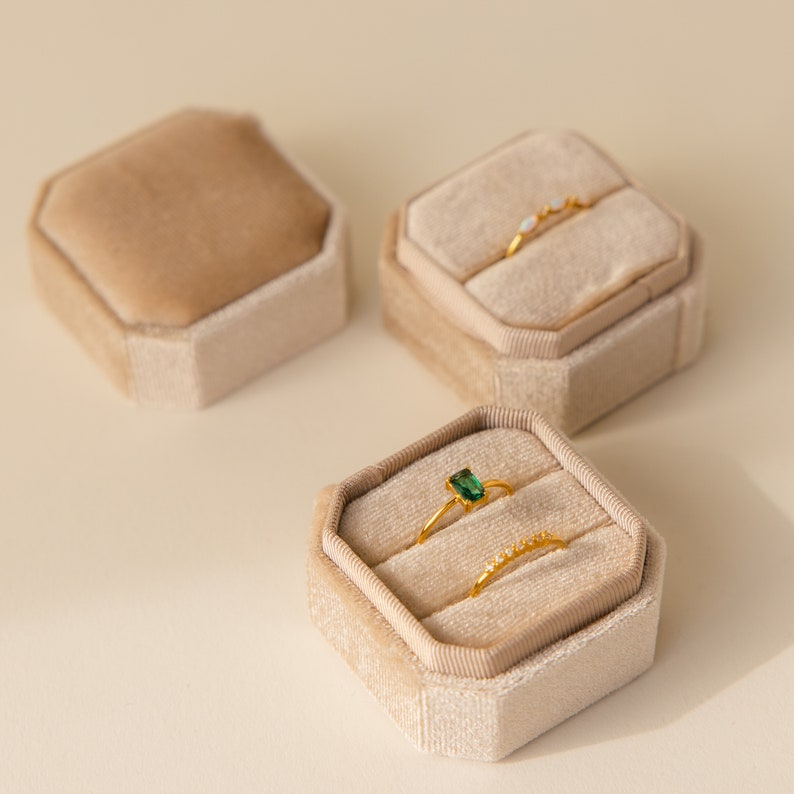 May include: Three velvet ring boxes in beige, with two rings inside one of the boxes. One ring has a green gemstone, and the other ring has small white stones.