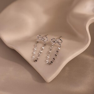 Bow Chain Stud Earrings in polished silver featuring delicate bow studs with a central stone and draped chain loops, displayed on a soft neutral surface.