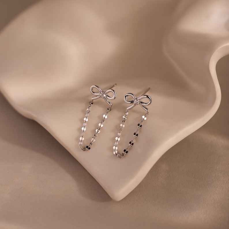 Bow Chain Stud Earrings in polished silver featuring delicate bow studs with a central stone and draped chain loops, displayed on a soft neutral surface.