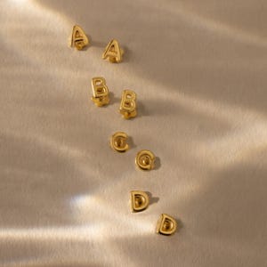 Gold initial stud earrings in pairs for the letters A, B, C, and D, arranged on a softly lit fabric surface with warm highlights and gentle shadows.