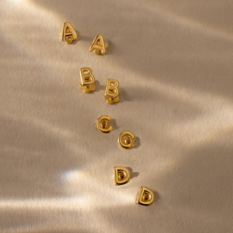 Gold initial stud earrings in pairs for the letters A, B, C, and D, arranged on a softly lit fabric surface with warm highlights and gentle shadows.