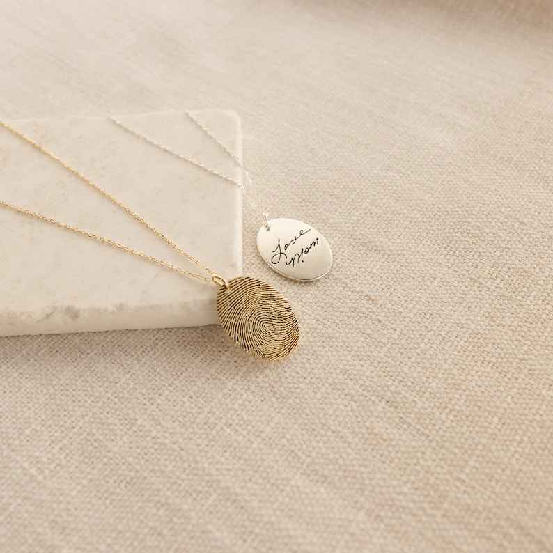 May include: Two silver necklaces with oval pendants. One pendant features a fingerprint design and the other has the handwritten text "Love Mom".