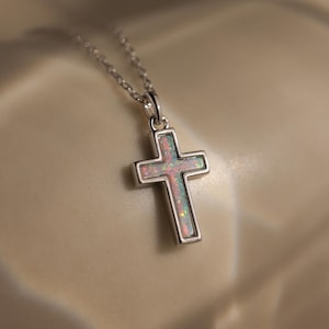 Könnte beinhalten: Eine silberfarbene Halskette mit einem Kreuzanhänger. Das Kreuz hat eine farbenfrohe, irisierende, opalartige Mitte, die von einem silbernen Rand umgeben ist. Die Kette ist zart und der Anhänger ist zentriert.
