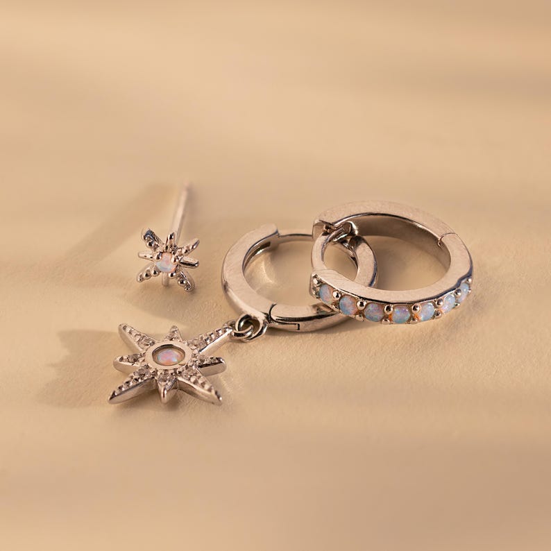 May include: A set of three silver earrings featuring a starburst design with opal accents. The earrings include a pair of hoop earrings and a single stud earring.