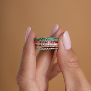 Peut inclure: Une pile de six bagues fines en or, chacune ornée d'une rangée de petites pierres précieuses serties pavé dans des couleurs distinctes, notamment vert, bleu, rose, transparent, violet et bleu clair, disposées en bandes fines.