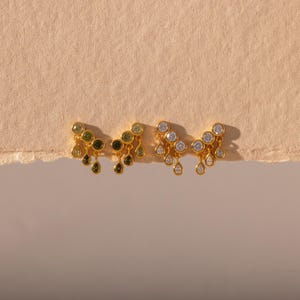 Puede incluir: Dos pendientes dorados con un conjunto de piedras engastadas. El pendiente izquierdo tiene piedras verdes en formas redondas y de lágrima, mientras que el derecho presenta piedras transparentes e incoloras en formas redondas y de lágrima.