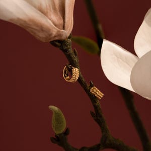 May include: A pair of gold-colored earrings, one a hoop and the other a stud, are displayed on a dark branch with a white flower and a burgundy background. The earrings have a textured, beaded design.