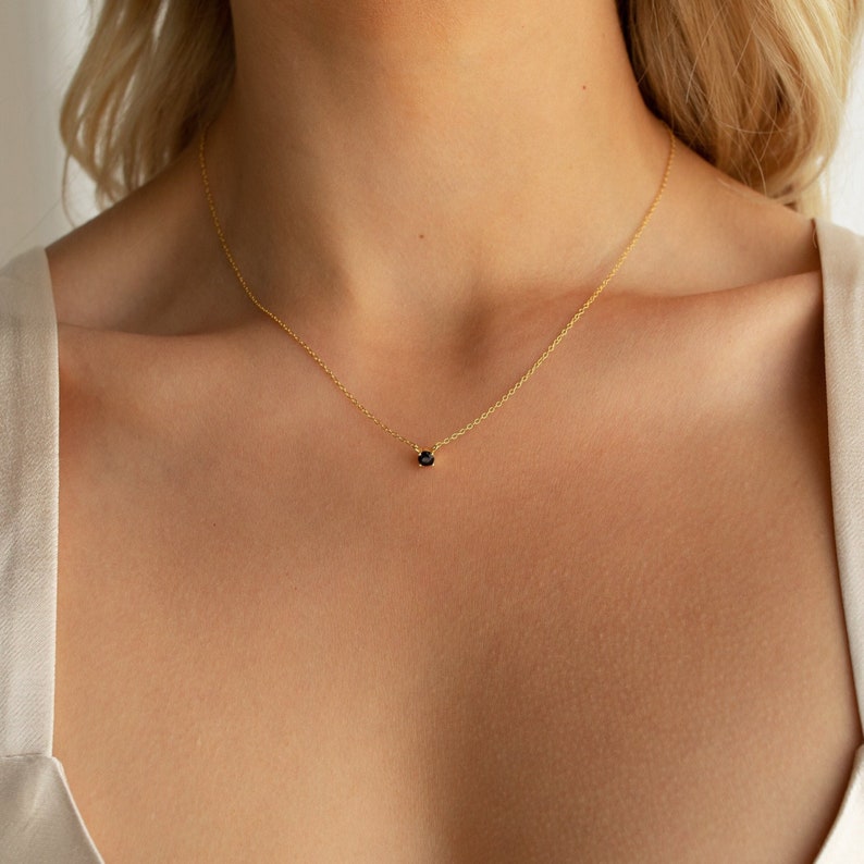 A close up of our Onyx Dainty Charm Necklace in 18K Gold finish on our model's neck hanging just along her collarbone - featuring a small round 4mm black CZ Diamond Charm centered on a dainty cable chain.