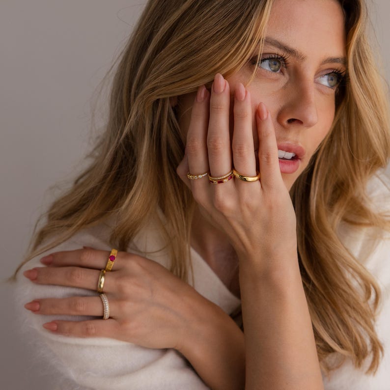 May include: A collection of gold rings, some with gemstones, displayed on a person's fingers. The rings vary in design, including a band with small diamonds, a band with red stones, and a simple gold band. The rings are set against a soft, neutral background.