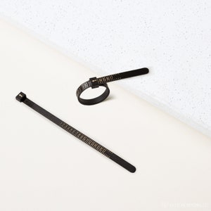 May include: Two black ring sizers with gold markings. One is a ring, and the other is a flat strip. Both have numbers indicating ring sizes. The ring sizer is on a white surface.