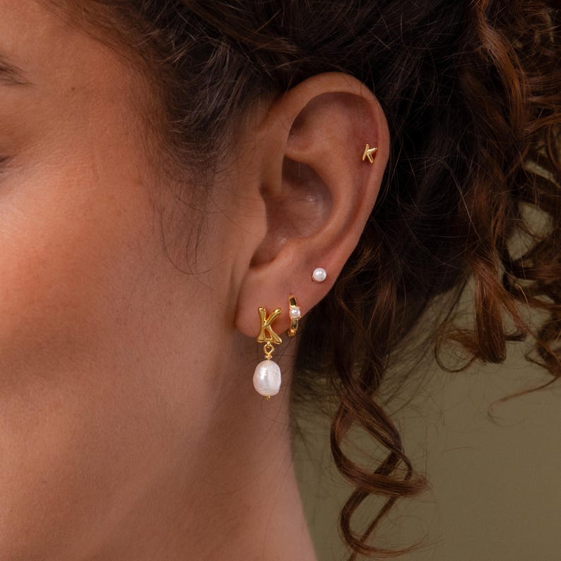 Gold bubble initial K stud earring styled in a layered ear stack, paired with a pearl drop charm and small pearl studs, shown on a model with curly hair.