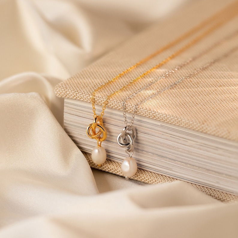 May include: Two necklaces with knot pendants and a single pearl drop. One necklace is gold-toned, the other is silver-toned. The necklaces are displayed on a book and a white satin cloth.