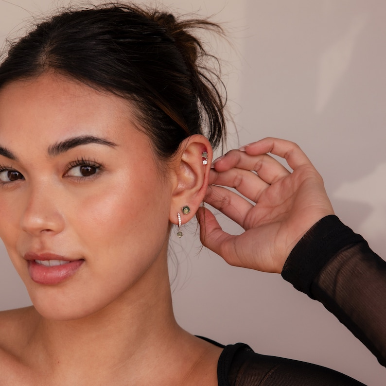 May include: A woman wearing a black shirt with a green gemstone earring and a silver stud earring.