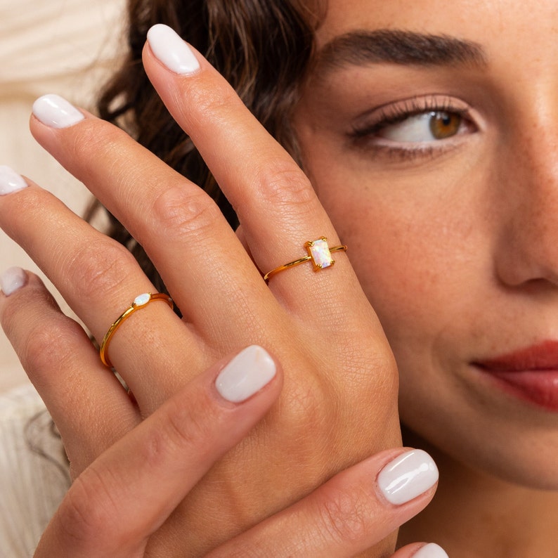 May include: Two gold rings with opal stones. One ring features a small oval opal, while the other has a rectangular opal. The rings are set against a soft, neutral background, highlighting the jewelry's delicate design and color.