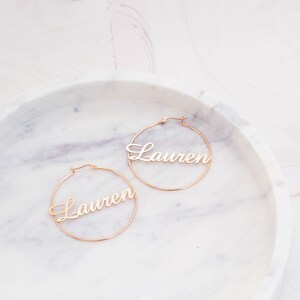 May include: A pair of gold hoop earrings with the name "Lauren" written in cursive on each hoop.