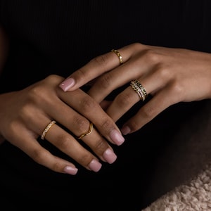 May include: Several gold rings of various designs are displayed on a person's hands. One ring has a row of small stones, another is a simple wave shape, and others have intricate details. The rings are set against a dark background.