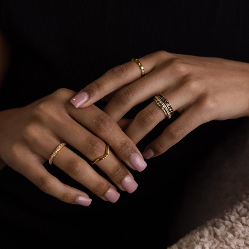 May include: Several gold rings of various designs are displayed on a person's hands. One ring has a row of small stones, another is a simple wave shape, and others have intricate details. The rings are set against a dark background.