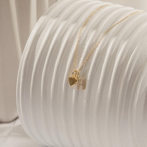 Puede incluir: Un delicado collar de oro con un colgante en forma de corazón y la letra "H" adornada con pequeños cristales. El collar se exhibe sobre una superficie blanca texturizada, mostrando su elegante diseño.