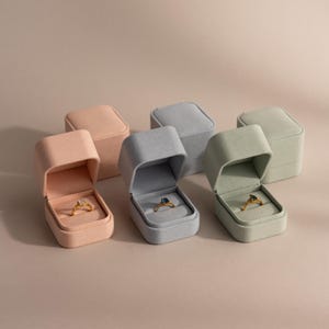 May include: Six velvet ring boxes in various pastel colors: peach, gray, and green. Three boxes are open, displaying gold rings with gemstones. The boxes are arranged on a neutral background, showcasing the jewelry.