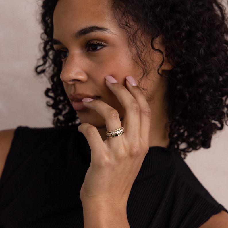 May include: A stack of gold rings with intricate detailing. The rings feature a mix of textures and designs, including what appears to be small gemstones. The rings are worn on a finger, showcasing the jewelry's craftsmanship and elegance.
