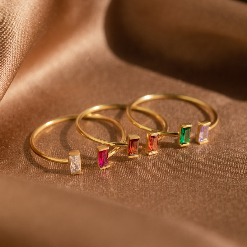 A close up of 3 of our Vertical Baguette Birthstone Rings in 18K Gold finish placed on a brown silk cloth - featuring 2.5mm x 5mm CZ Gemstones, one ring with clear crystal and ruby, one with 2 Garnets, and the last with emerald and clear crystal