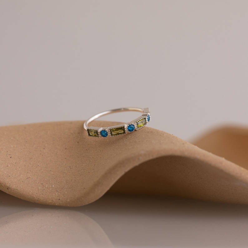 May include: A silver ring with a row of alternating gemstones. The ring features round and rectangular stones in shades of blue, green, and yellow. The ring is set on a neutral background.