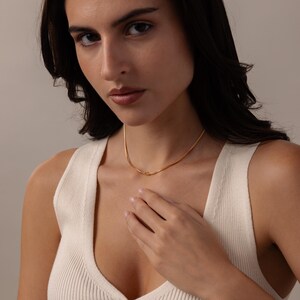 May include: A delicate gold necklace with a subtle knot design. The necklace is displayed on a person wearing a white tank top. The gold chain is thin and appears to be made of small, interconnected links.