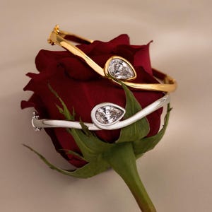 May include: Two hinged bangle bracelets, one in polished gold tone and one in silver tone, each featuring a single pear-shaped clear gemstone set at the center. The bracelets are resting on a deep red rose with green sepals and a stem.