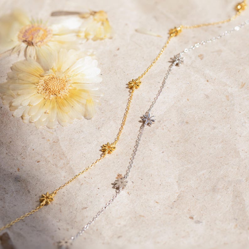 May include: Two delicate necklaces with small flower charms. One necklace is gold and the other is silver. The necklaces are laying on a textured beige surface.
