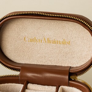 May include: A brown leather jewelry box with a gold zipper. The inside of the box is lined with a soft, beige fabric. The box has the words 'Caitlyn Minimalist' embossed in gold on the inside.