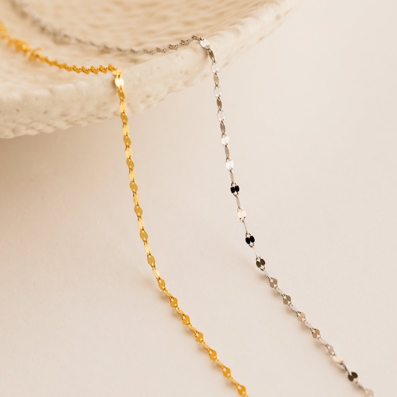 A close up of 2 of our Josephine Oval Chain Bracelets, one in the gold finish and the other in silver, dangling off the edge of a textured cream white clay jewelry dish - featuring dainty mirror link twist chains.