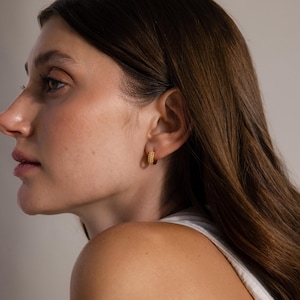 May include: A close-up shot of a person wearing a pair of small, gold hoop earrings. The earrings have a textured, beaded design. The person has long, brown hair and is wearing a white tank top.
