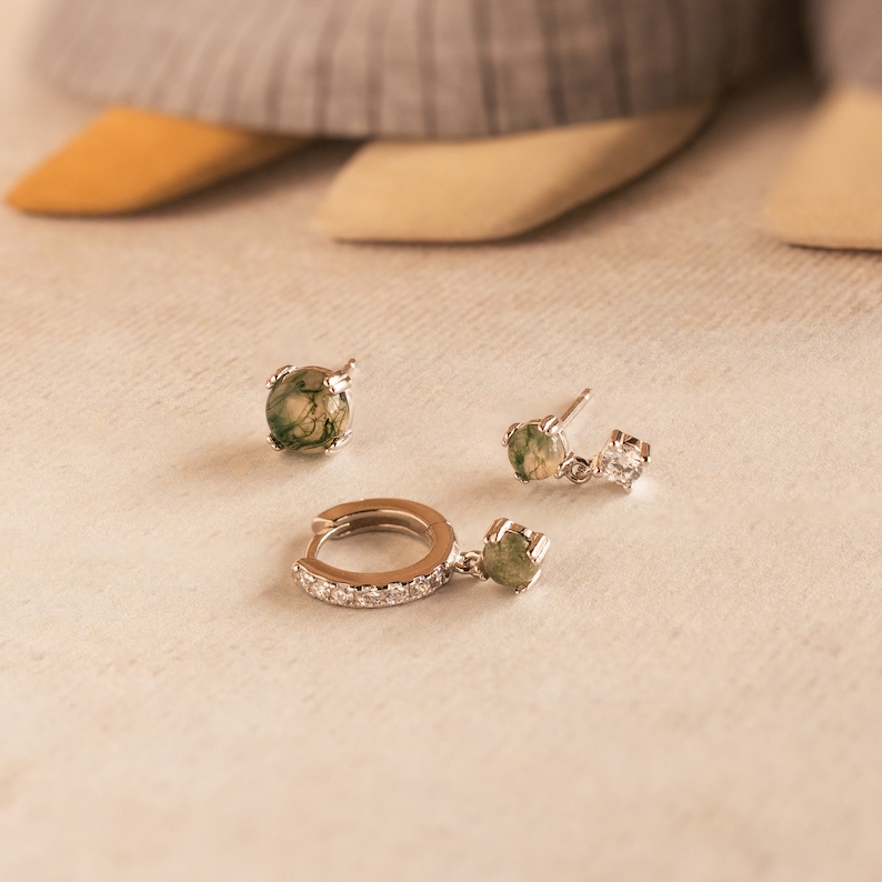 May include: A set of three silver earrings with green and white stones. The earrings are on a white surface.