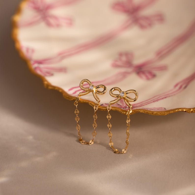 Bow Chain Stud Earrings in gold featuring delicate bow studs with a central stone and draped chain loops, displayed on a decorative scalloped dish with pink ribbon design.
