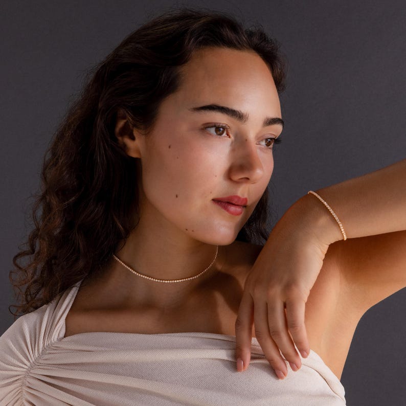 May include: A delicate gold beaded necklace and bracelet set. The necklace is a choker style, and the bracelet is a simple strand. Both pieces are made of small, round beads. The jewelry is displayed on a person with fair skin and dark hair.