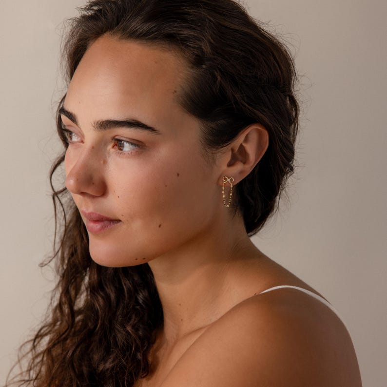 Woman wearing Bow Chain Stud Earrings in gold with delicate bow studs and draped chain loops, styled with minimal makeup and natural waves.