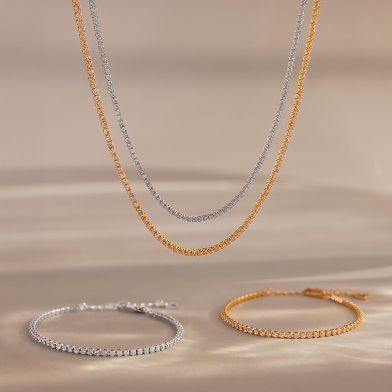 May include: Two necklaces and two bracelets. One necklace and bracelet are gold-toned, the other necklace and bracelet are silver-toned. All four pieces are delicate, with small, evenly spaced embellishments. The jewelry is displayed on a neutral background.