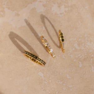 May include: Four gold rings with various gemstone arrangements. One ring features dark green stones, another has white stones, and a third has a mix of green and white stones. The fourth ring has a full band of dark green stones.