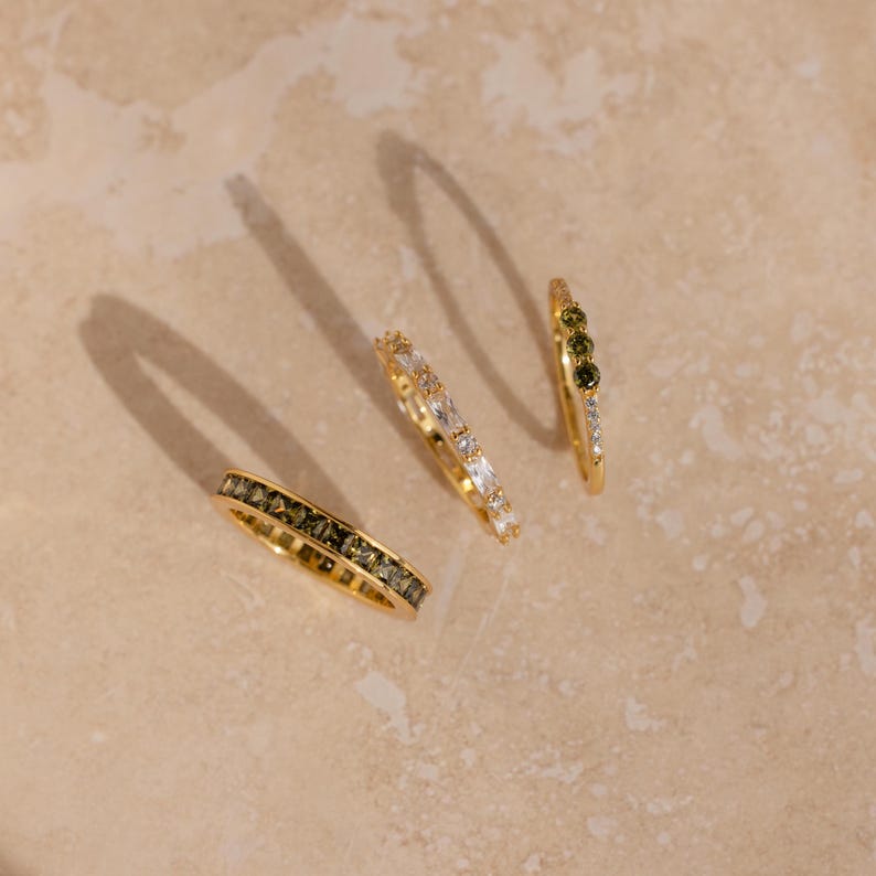 May include: Four gold rings with various gemstone arrangements. One ring features dark green stones, another has white stones, and a third has a mix of green and white stones. The fourth ring has a full band of dark green stones.