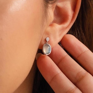 May include: A silver-toned earring featuring a small, round, clear stone at the top post, connected to a larger, smooth, oval-shaped, translucent grey stone drop. The earring is secured to the earlobe with a simple metal backing.