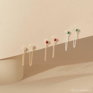May include: Three pairs of chain earrings featuring bezel-set stones. The first pair is gold with clear stones, the second is gold with red stones, and the third is silver with green stones. Each earring has a stud top and a dangling chain.