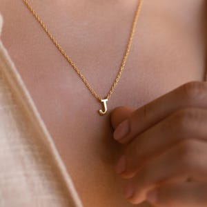 May include: A gold-colored necklace with a delicate chain and a pendant in the shape of the letter "J". The necklace is displayed on a person's chest, with the chain resting against the skin. The pendant is small and simple.