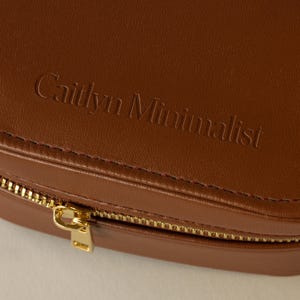 May include: A brown leather jewelry box with a gold zipper. The box has the words 'Caitlyn Minimalist' embossed on the top.