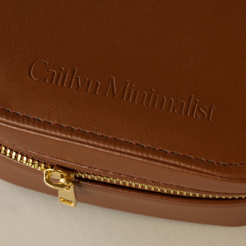 May include: A brown leather jewelry box with a gold zipper. The box has the words 'Caitlyn Minimalist' embossed on the top.