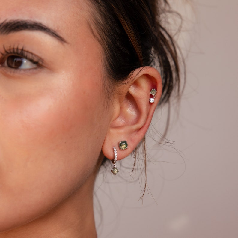 May include: A silver hoop earring with a small green gemstone dangling from the bottom. The hoop is set with small clear gemstones. The earring is worn in the lower lobe of a person's ear. The person is also wearing a silver stud earring with a small green gemstone in the upper lobe of the same ear.