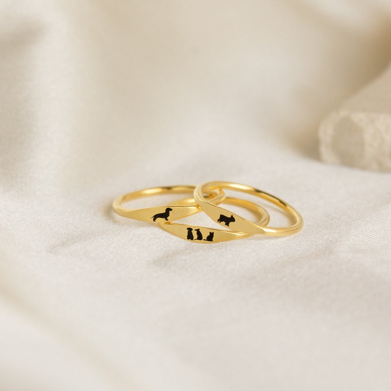 Three dainty gold rings each with an engraved pet design in black ink. The middle ring has a combination of three different pets, both dogs and cats, engrave on it which the other two have one dog engraved on each.