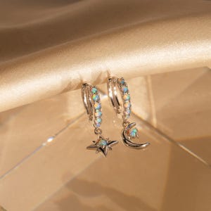 May include: A pair of silver-toned hoop earrings featuring small iridescent opal stones along the hoops. One earring has a dangling four-pointed star charm with a central opal, and the other has a crescent moon charm with a small opal accent.