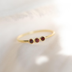 Pode incluir: Um anel fino de ouro com três pequenas pedras preciosas granadas redondas de cor vermelho-escura dispostas numa fila horizontal. A banda é lisa e metálica, apresentando um design delicado com as três pedras engastadas num aro simples.